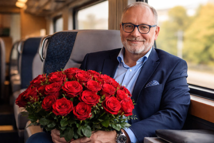Gepflegter älterer Mann mit Brille sitzt im Zug und hält einen großen Strauß roter Rosen für ein Date